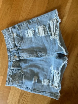Ljusa blå jeansshorts med slitningar - Säljer ett par högmidjade ljusblå jeansshorts med råa kanter och flera slitningar framtill. Klassisk femficksmodell med knapp och dragkedja. Perfekta för sommaren och ger en avslappnad streetstyle-vibe. 💕 pris kan diskuteras 