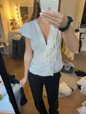 Vit prickig blus med v-ringning - Snygg vit blus med svarta prickar och djup v-ringning. Blusen har korta, fladdriga ärmar och markerad midja som ger en feminin siluett. Perfekt att styla med både jeans och kostymbyxor för en trendig look. Vet ej storlek eller märke men passar mig som är en S!