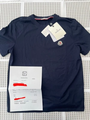 Mörkblå Moncler t-shirt i bomull - Snygg mörkblå t-shirt från Moncler med klassisk rund hals och korta ärmar. T-shirten har Monclers ikoniska logotyp broderad på bröstet och diskreta detaljer vid halsringningen. Tillverkad i mjuk bomull för en skön känsla.