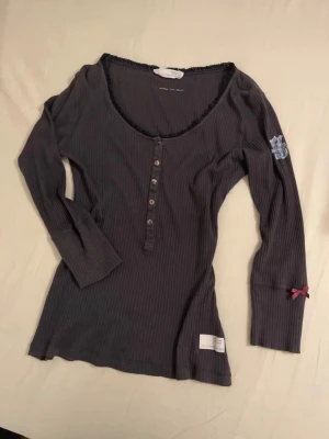 Grå ribbad topp Odd Molly - Mörkgrå långärmad ribbad topp från Odd Molly med knappar framtill, spetskant vid halsen. Har sytt in 1 cm på sidorna, men inget som syns. Tröjan är stretchig så passar XS-S.