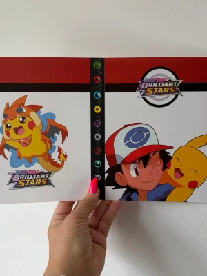 Pokémon Brilliant Stars samlarpärm - Säljer en Pokémon Brilliant Stars samlarpärm med färgglada motiv av Pikachu och Ash på framsidan. Pärmen är vit och röd med plastfickor inuti för att förvara samlarkort. Perfekt för Pokémonkort och har tydliga symboler för olika element på ryggen.