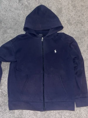 Mörkblå hoodie från Polo Ralph Lauren - Snygg mörkblå hoodie från Polo Ralph Lauren med dragkedja och klassisk logga broderad på bröstet. Hoodien har huva, två fickor framtill och ribbade muddar. Perfekt för en avslappnad och cool look. Den här blivet lite lila och saknar snören, har gärna av er vid minsta fundering✅