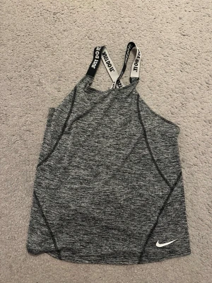 Grå linnetopp från Nike - Snygg gråmelerad linnetopp från Nike med svarta och vita axelband där det står 'JUST DO IT'. Toppen har en sportig look och är perfekt för träning. Materialet känns mjukt och stretchigt, och modellen är ärmlös med korsade band i ryggen. Passar bra för ex en 9-12 åring. 