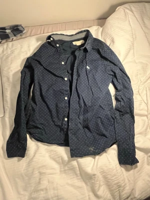 Blå prickig skjorta från L.O.G.G. - Mörkblå skjorta från L.O.G.G. med vita prickar och klassisk krage. Skjortan har långa ärmar och knäppning framtill med vita knappar. Tillverkad i bomull, perfekt för en avslappnad men ändå stilren look.