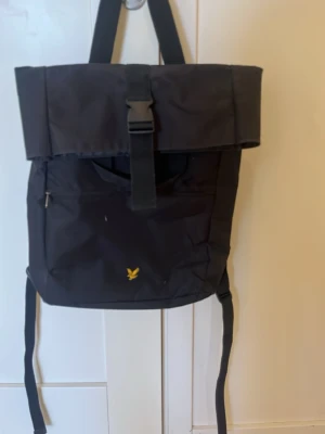 Svart ryggsäck från Lyle & Scott - Snygg svart ryggsäck från Lyle & Scott 45*40cm,  med gul logga framtill. Ryggsäcken har ett stort lock med spänne, justerbara axelband och en dragkedjeficka på framsidan. Även plats f pc med dragkedja bak. Perfekt för skolan eller stan, rymmer mycket och har stilren design. Rök- och djurfritt hem. 