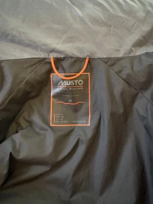 Grå jacka från Musto Land Rover - Säljer en grå pufferjacka från Musto Land Rover i storlek M. Jackan har hög krage, hel dragkedja och quiltad design. Invikbar luva.Perfekt för kalla dagar, med tekniskt material som håller värmen. Diskret logga på bröstet och orange detaljer vid dragkedjan.