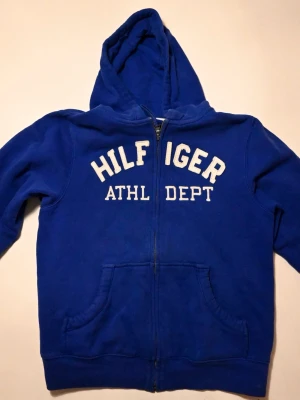 Blå hoodie från Tommy Hilfiger - Snygg blå hoodie från Tommy Hilfiger med dragkedja framtill och vit broderad text 'HILFIGER ATHL DEPT' på bröstet. Utrustad med stor huva och två fickor framtill. Perfekt för en avslappnad och sportig stil.