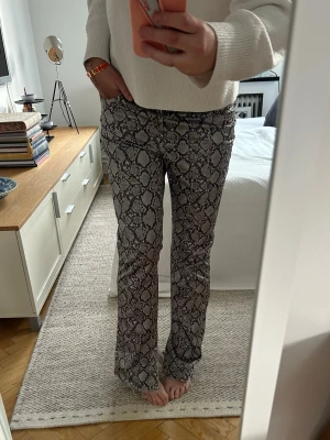 Bootcut jeans med ormmönster - Säljer ett par snygga bootcut byxor med grått och svart ormmönster. Jeansen har normal midja och är i stretchigt material för skön passform. Perfekta för dig som vill sticka ut med ett trendigt mönster. Med slits, passar ca M❤️