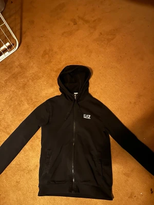 Svart hoodie från EA7 Emporio Armani - Säljer en svart hoodie från EA7 Emporio Armani med dragkedja och två fickor framtill. Hoodien har en klassisk huva med snören och logotyp på bröstet. Materialet är mjukt och bekvämt, perfekt för en avslappnad stil. ”Storlek S”