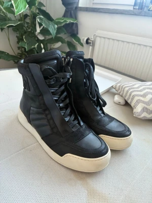 Svarta high top sneakers från Bally - Snygga svarta high top sneakers från Bally med vita sulor, dragkedja på sidan och breda skosnören. Skorna har en cool design med detaljer i både läder och mocka samt Bally-logga broderad på hälen. Perfekta för dig som gillar streetwear och vill sticka ut.
