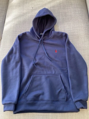 Marinblå hoodie från Polo Ralph Lauren - Snygg marinblå hoodie från Polo Ralph Lauren med klassisk röd logga broderad på bröstet. Hoodien har huva med dragsko, stor magficka och ribbade muddar. Tillverkad i mjuk bomullsblandning som känns skön mot huden. Perfekt för en avslappnad och stilren look.