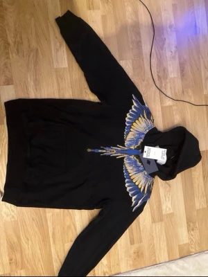 Svart hoodie från Marcelo Burlon - Säljer en svart hoodie från Marcelo Burlon med ikoniskt färgstarkt vingmönster i blått, gult och vitt över axlar och bröst. Hoodien har huva och känguruficka framtill. Materialet är mjukt och bekvämt, perfekt för en streetwear-look.