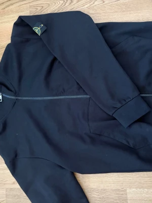 Svart hoodie från Stone Island - Säljer en svart hoodie från Stone Island med dragkedja framtill och klassisk logopatch på ärmen. Hoodien har ribbade muddar och fickor på sidorna. Tillverkad i mjuk bomull för en skön och avslappnad look.