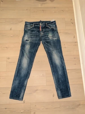 Mörkblå Dsquared2 jeans med slitningar - Säljer ett par mörkblå jeans från Dsquared2 i storlek 52. Jeansen har en slim fit passform, slitningar, färgstänk och en tvättad look som ger en cool och trendig vibe. Perfekta för dig som gillar streetwear och vill sticka ut lite extra.