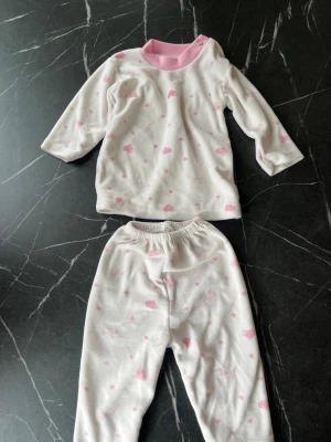 Vit och rosa pyjamas med hjärtan - Söt tvådelad pyjamas i mjuk vit trikå med rosa hjärtan och rosa mudd vid halsen. Tröjan har långa ärmar och tryckknapp vid axeln för enkel påklädning. Byxorna har resår i midjan för skön passform. Perfekt för mysiga kvällar hemma.