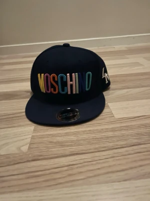 Moschino keps blå - Snygg marinblå keps från Moschino med broderad logga i regnbågens färger fram och vita bokstäver på sidan. Klistermärket sitter även kvar där fram 