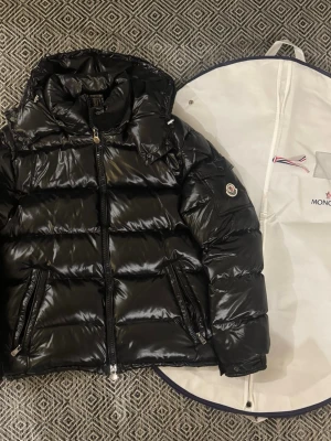 Svart dunjacka från Moncler - Snygg svart dunjacka från Moncler med glansig finish och klassisk pufferdesign. Jackan har huva, två dragkedjeförsedda fickor och ikonisk Moncler-logga på ärmen. Insidan har ett serietidningsmönster och jackan levereras med original klädpåse.