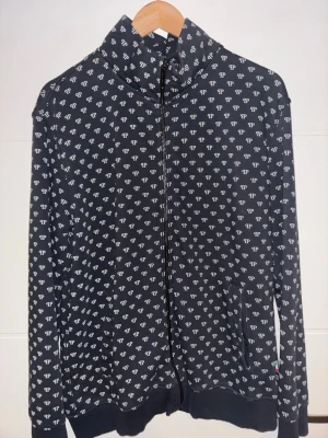 Svart hoodie från Philipp Plein - Cool svart hoodie från Philipp Plein med vitt all-over logomönster och dragkedja framtill. Tröjan har hög krage, ribbade muddar och fickor på sidorna. Materialet är mjukt och bekvämt, perfekt för en avslappnad streetstyle-look.