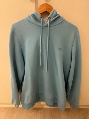 Ljusblå hoodie från HUGO - Snygg ljusblå hoodie från HUGO med minimalistisk design och diskret logga på bröstet. Hoodien har huva med långa dragsnören och ribbade muddar vid ärmslut och nederkant. Tillverkad i mjukt bomullsmaterial som känns skönt mot huden.