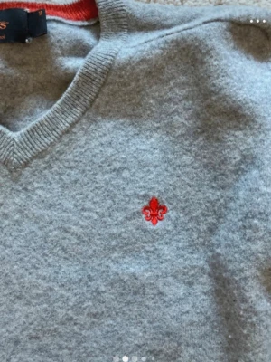 Ljusgrå stickad tröja med röd detalj - Ljusgrå stickad tröja från Morris med v-ringning och lång ärm. Tröjan har en liten röd broderad lilja på bröstet och ribbade muddar vid ärmslut och nederkant. Perfekt för lager på lager och enkel att matcha med jeans eller kjol.