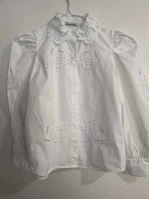 Vit blus med broderad krage från Zara - Söt vit blus från Zara med puffiga långärmar och detaljerade broderier på både fram- och baksida. Blusen har en stor, spetsbroderad krage och knäppning framtill. Materialet är bomull och ger en luftig känsla. Perfekt för dig som gillar romantisk och trendig stil.