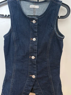 Ärmlös jeansväst  - Snygg ärmlös jeansväst i mörkblå denim från Perfect Jeans. Västen har rund halsringning, knäppning framtill med silverfärgade knappar och markerade sömmar för en cool look. Perfekt att styla över en t-shirt eller tröja.