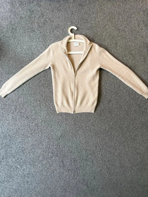 Beige Cashmere John Henric Tröja - Snygg beige ziptröja med ribbad struktur och hög krage. Tröjan har långa ärmar och dragkedja framtill, perfekt för lager på lager. Tillverkad i mjukt material som känns skönt mot huden.