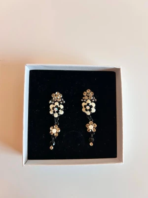 Blommiga örhängen med pärldetaljer - Säljer ett par snygga örhängen med vita och svarta blommor, dekorerade med små glittrande stenar och pärlliknande detaljer. Örhängena har en hängande design och ger en cool och unik look. Perfekta för dig som gillar statement-smycken och vill sticka ut.