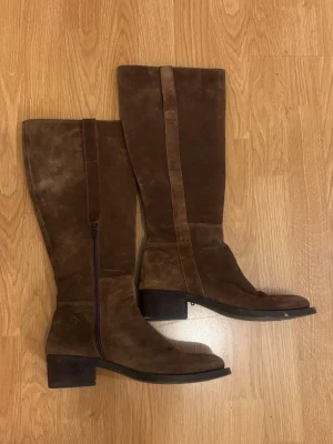 Bruna knähöga mockastövlar - Snygga bruna knähöga stövlar i mocka med bred klack och dragkedja på insidan. Klassisk design med rundad tå och robust sula. Perfekta till höstens outfits och ger en stilren look.