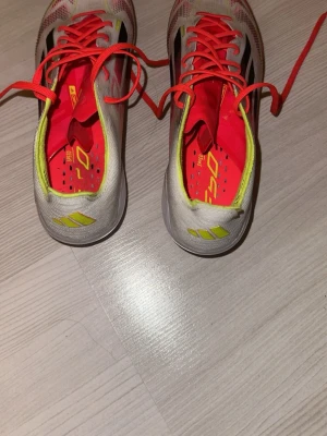  Adidas  f50 elite  - Säljer ett par Adidas f50 fotbollsskor i vitt med svarta och röda detaljer, samt neongula inslag och röda snören. Skorna har en lätt och smidig design med FG-dobbar för spel på naturgräs. Ovandelen är tillverkad i syntetmaterial för bra bollkontroll.
