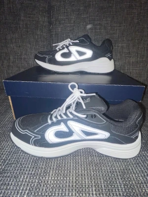 Svarta dior sneakers med vit detalj - Snygga svarta sneakers från Christian Dior med vita reflekterande detaljer och tjock vit sula. Skorna har en sportig design med mesh och syntetmaterial, samt vita skosnören. Perfekta för dig som gillar streetwear och vill sticka ut.