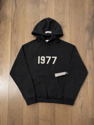 Svart 1977 hoodie från Essentials - Snygg svart hoodie från Essentials med trycket '1977' i vitt på bröstet. Klassisk känguruficka framtill och huva. Tillverkad i mjukt bomullsmaterial som känns skönt mot huden. Perfekt för en avslappnad och trendig look.