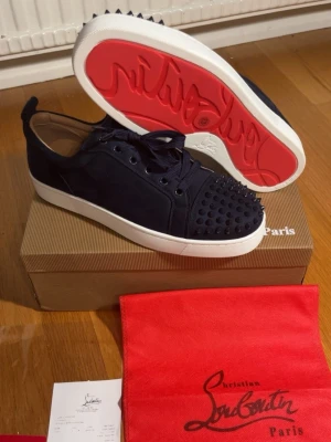 Svarta Louboutin sneakers med nitar - Snygga svarta sneakers från Christian Louboutin med mattsvarta nitar på tån och klassisk röd sula. Skorna har en låg profil, vita sulor och är tillverkade i mocka med läderdetaljer. Perfekta för dig som vill sticka ut med exklusiv design och edgy detaljer.