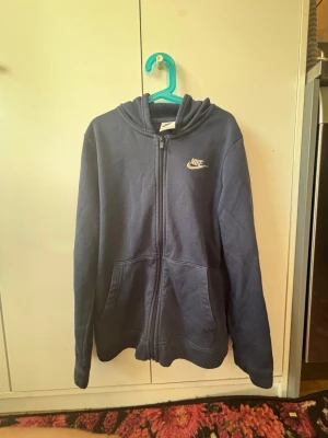 Mörkblå zip hoodie från Nike - Snygg mörkblå hoodie från Nike med dragkedja framtill och klassisk vit Nike-logga broderad på bröstet. Hoodien har huva och två stora fickor framtill. Tillverkad i mjukt bomullsmaterial som är skönt att ha på sig.