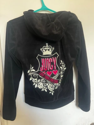 Svart velour hoodie från Juicy Couture - Säljer en svart velour hoodie från Juicy Couture med glittrande stenar och broderade detaljer. Stor Juicy-text framtill och ett stort emblem med krona, hjärtan och blommor på ryggen. Dragkedja och kängurufickor gör den både snygg och praktisk. Perfekt för en trendig och avslappnad look.