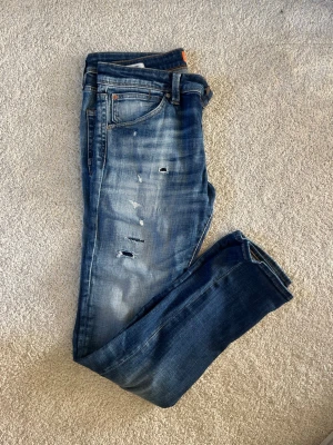 Slim Glenn blå slitna jeans - Säljer ett par blå slim fit jeans från Slim Glenn med slitningar och distressed detaljer på framsidan. Jeansen har klassisk femficksmodell, normal midja och är tillverkade i denim med snygg tvättad look. Perfekta för en avslappnad och trendig stil.