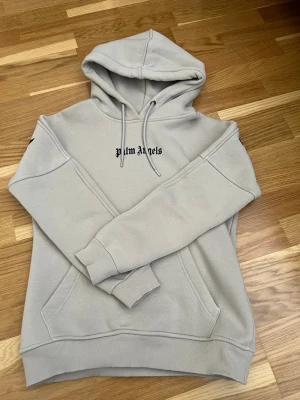 Beige hoodie från Palm Angels - Snygg beige hoodie från Palm Angels med svart logga broderad på bröstet. Klassisk huva med dragsko och magficka framtill. Tillverkad i mjukt bomullsmaterial och har ribbade muddar vid ärmslut och nederkant. Perfekt för en avslappnad streetwear-look. Det finns en liten fläck vid halskragen där snöret sitter - se bild 4. 