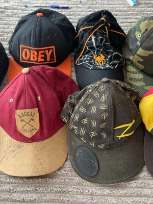 Samling kepsar från olika märken (25kr styck) - Säljer ett gäng kepsar i olika stilar och färger, bland annat en svart och orange Obey-keps, en vinröd och beige Rackartygarna-keps, en svart keps med spindelmotiv, en kamouflagekeps och en färgglad Ice Hockey World Championship 95-keps. Perfekt för dig som vill variera din stil!