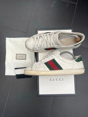 Gucci Ace Sneakers – Original med box & dustbag - Säljer ett par klassiska Gucci Ace sneakers i vitt läder med den ikoniska gröna/röda web-randen. En tidlös modell som fungerar till både jeans, chinos och mer uppklädda outfits.  Skorna är använda och har normalt slitage i form av veck på lädret samt bruksspår på sulor och tåparti, vilket framgår av bilderna. Fortfarande mycket snygga på fot och har många år kvar.                                    ✅Originalbox medföljer ✅ Dustbag medföljer ✅ Skoblock medföljer ✅ Klassisk och eftertraktad modell