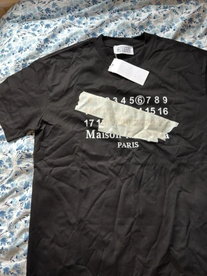 Maison Margiela svart t-shirt - Svart t-shirt från Maison Margiela med ikoniskt tryck framtill i vitt och beige, samt texten 'Maison Margiela PARIS'. Klassisk rund hals och korta ärmar. Tillverkad i mjuk bomull för en bekväm känsla. Perfekt för dig som gillar streetwear och designerplagg. Helt ny storlek M