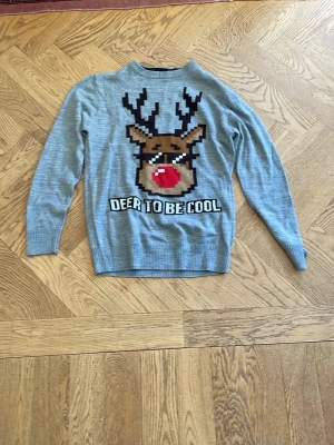 Tjocktröjor - Grå stickad tröja med ett stort motiv av en ren med solglasögon och röd nos på framsidan. Texten 'DEER TO BE COOL' under motivet. Perfekt för julen och har ribbade muddar vid ärmslut och nederkant.                                                                Båda storlek 146-152