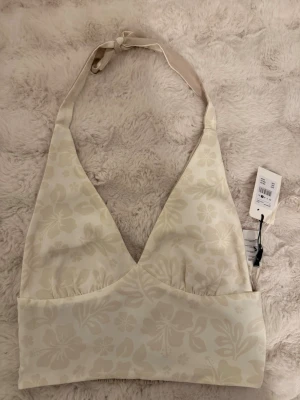 Beige blommig halterneck linnetopp - Säljer en beige halterneck linnetopp från Bik Bok med subtilt blommönster.  Toppen är helt ny då den även har prislapp kvar. Toppen knyts i nacken och har en bred resår under bysten för extra komfort. Perfekt för varma dagar och somriga outfits.
