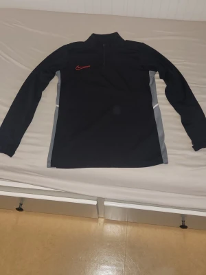 Svart Nike träningströja med dragkedja - Säljer en svart långärmad träningströja från Nike med halv dragkedja framtill och röd Nike-logga på bröstet. Tröjan har gråa paneler längs sidorna och är tillverkad i ett stretchigt funktionsmaterial som passar perfekt till träning eller uppvärmning. PRISET ÄR INTE HUGGET I STEN!!🔥🤝 