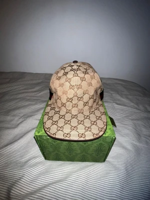 Gucci keps  - Säljer en beige keps från Gucci med det klassiska GG-monogrammet över hela kepsen. Kepsen har en böjd skärm och en grön-röd rand på sidan. Helt ny pris kan diskuteras 