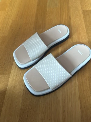 Beige sandaler från BOSS med mönster - Snygga beige sandaler från BOSS med bred rem i textil och diskret prickigt mönster. Sandalerna har fyrkantig tå och vit sula med svart undersida. Perfekta för en avslappnad och trendig look.