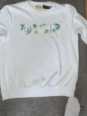 Vit sweatshirt  - Säljer en vit sweatshirt  med stort tryck i grönt och vitt på bröstet. Tröjan har rund halsringning, ribbade muddar vid ärmslut och nederkant samt en clean och stilren look. Perfekt för dig som gillar exklusiva märken och enkel design.