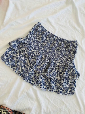 Blommig kortkjol med volang - Supersöt kortkjol med blått blommönster på vit botten. Kjolen har smock i midjan och volangdetaljer som ger en flowy känsla. Perfekt för varma dagar och enkel att styla med en enkel topp. Skriv för fler bilder🥰