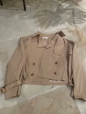 Kort beige trenchcoat med bälte - Trendig kort trenchcoat i beige. Jackan har dubbelknäppning framtill, bred krage och bälte vid ärmsluten för en cool look. Cropped passform och minimalistisk design gör den enkel att styla. 