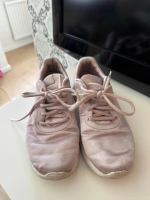 Nike sneakers ljusrosa - Säljer ett par ljusrosa Nike sneakers med klassisk låg profil och snörning. Skorna har en vit, tjock sula och är tillverkade i ett glansigt tygmaterial. Swoosh-loggan syns på sidan för en clean look. Perfekta för dig som gillar sportig och stilren vibe.