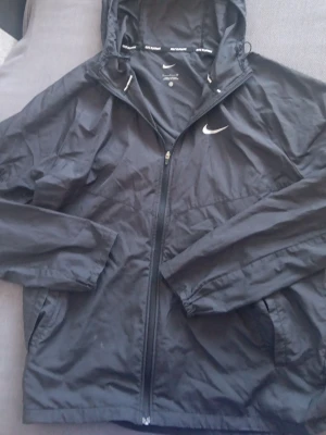 Svart Nike vindjacka med huva - Snygg svart vindjacka från Nike med dragkedja framtill och klassisk vit Nike-logga på bröstet. Jackan har huva med dragsko och två sidofickor. Tillverkad i lätt och tåligt material som passar perfekt för blåsiga dagar.